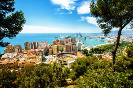 Málaga, Spanien, mit der Stierkampfarena La Malagueta im Vordergrund, umgeben von Gebäuden und Bäumen. Im Hintergrund erstreckt sich der Hafen mit Schiffen und Kränen bis zum Mittelmeer unter einem blauen Himmel mit einigen Wolken
