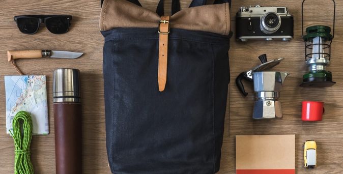 Verschiedene Reiseutensilien wie Rucksack, Sonnenbrille und Kamera liegen auf einem Holztisch.