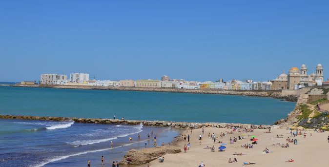 Blick über Strand mit Badegästen, das Meer und die Silhouette von Cadiz. Die Kathedrale sticht hervor. Blauer Himmel und kleine Wellen im Meer