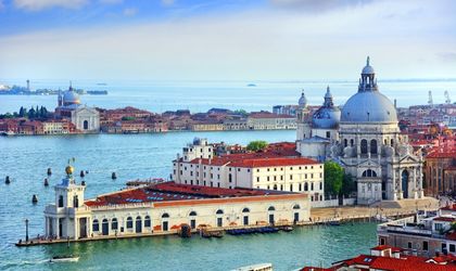 Die imposante Basilika Santa Maria della Salute in Venedig, Italien, mit ihrer markanten Kuppel und den venezianischen Gebäuden am Canal Grande, unter einem blauen Himmel.   Sources