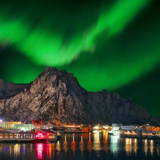 Die atemberaubenden Nordlichter am Himmel. Darunter ein Berg mit kleinen beleuchteten Häusern.