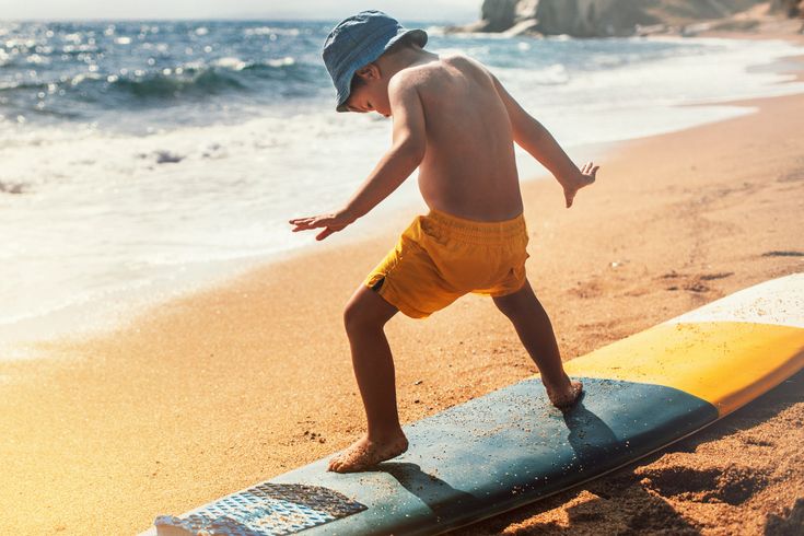 Ein kleiner Junge mit orangener Badehose steht auf einem Surfboard, welches noch am Starnd liegt.