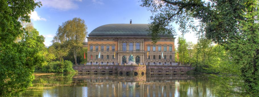 Eine ruhige Szene des Kaiserteichs im Ständehaus Park in Düsseldorf, Deutschland, mit der Kunstsammlung Nordrhein-Westfalen am Ufer, umgeben von üppigem Grün und einem klaren Himmel.