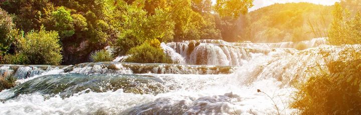 Eine Landschaft mit einem Wasserfall und Gebäuden bei Sonnenuntergang.