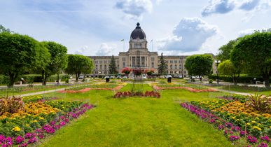 Das Parlamentsgebäude von Saskatchewan in der Hauptstadt Regina. Davor eine weitläufige Wiese, viele angelegte farbenfroh blühenden Blumen, Bäume. Parkähnlich angelegt. Einige Autos parken vor dem Gebäude. Das alles bei blauem Himmel.