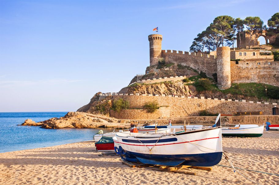 Ein Fischerboot liegt am Sandstrand vor der mittelalterlichen Festung Tossa de Mar an der Costa Brava.