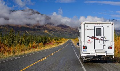 Camper von hinten zu sehen, welcher auf der Straße fährt. Umgeben von Wäldern. Blauer Himmel.