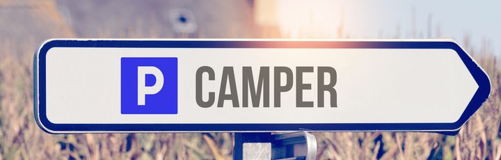 Blaues Schild mit weißem Pfeil und dem Symbol "P Camper" auf einem Schotterweg.