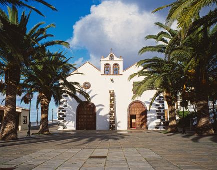 Altstadt Santo Domingo: ine weiße Kirche mit einem dunklen Dach und einem Glockenturm, umgeben von hohen Palmen und einem gepflasterten Vorplatz unter einem teilweise bewölkten blauen Himmel.