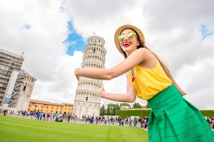 Der Schiefe Turm von Pisa im Hintergrund. Im Vordergrund, sehr groß abgebildet, eine junge Frau mit grünem Rock und gelben Shirt, Sonnenbrille und Hut. Sie umfasst den Turm.