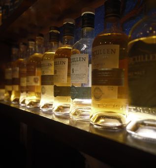 Eine Bar mit einer Reihe von beleuchteten Whiskeyflaschen, die auf einem Regal stehen. Die Flaschen sind unterschiedlich groß und geformt und zeigen verschiedene Whiskeymarken.