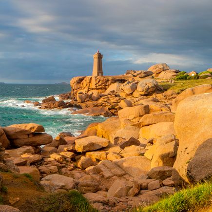 Eine beeindruckende felsige Küstenlandschaft mit rosafarbenen Granitfelsen, die sich ins Meer erstrecken. Der Sonnenuntergang taucht die Szenerie in warme Orangetöne und schafft eine dramatische Atmosphäre.