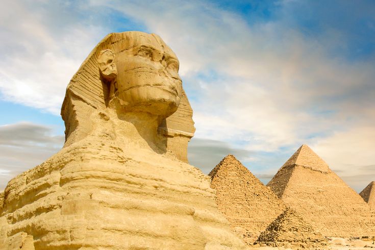 Eine Nahaufnahme der Großen Sphinx von Gizeh, mit zwei der Pyramiden von Gizeh im Hintergrund unter einem bewölkten Himmel.