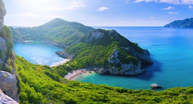 Die Doppelbucht von Porto Timoni auf Korfu, Griechenland, mit zwei Stränden, die von üppiger grüner Vegetation und steilen Klippen umgeben sind, während das blaue Meer im Hintergrund liegt.