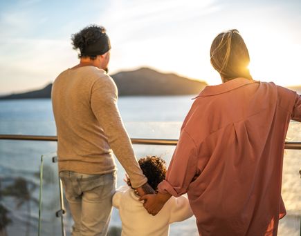 PAar steht mit Kind an der Reeling eines Kreuzfahrtschiffes und blickt auf das Meer