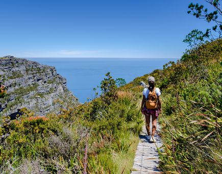 Eine Frau mit kurzer Hose und Rucksack wandert einen Trampelpfad oberhalb der Küste. Rundherum alles grün und hellblauer Himmel und klares Meer.
