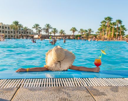 Frau mit Sonnenhut im Hotelpool, in der rechten Hand ein Cocktail. Im Hintergrund die Hotelkulisse.