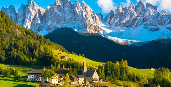Die Dolomiten. Im Vordergrund grüne Wiese mit kleinem urigen Dorf, direkt am Waldhang. Dahinter die prächtigen, schneebedeckten Dolomiten bei blauem Himmel.