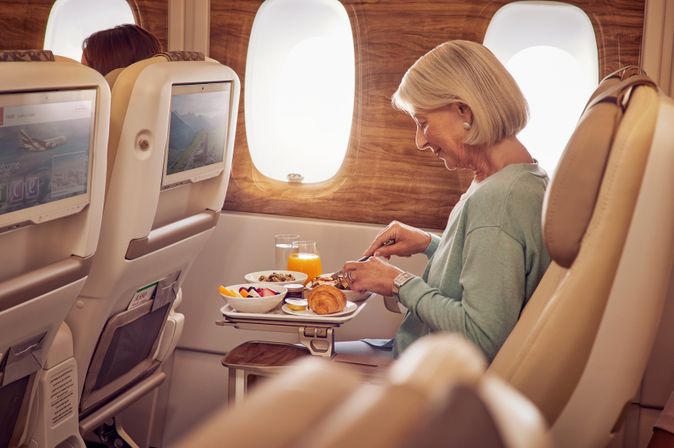 Passagierin frühstückt an Bord mit Croissant, Obst und Getränken auf einem ausklappbaren Tisch vor dem Bildschirm.