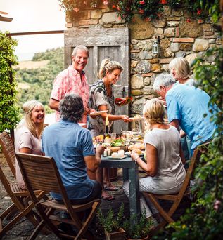 Eine Familie sitzt an einem Tisch im Freien und genießt eine Mahlzeit in einer malerischen Umgebung, umgeben von Bäumen und einem warmen Licht.