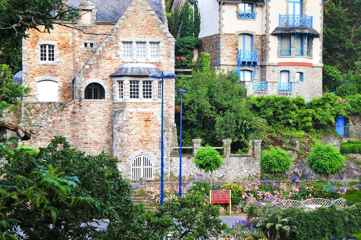 Ein charmantes, altes Steinhaus mit einer traditionellen Architektur, umgeben von grünen Pflanzen und einem blauen Himmel. Das Haus wirkt sehr malerisch.