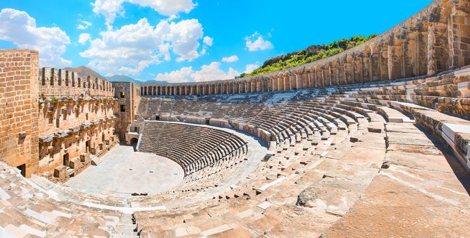 Das antike römische Theater von Aspendos in der Türkei, ein beeindruckendes, gut erhaltenes Amphitheater mit vielen Sitzreihen und einer großen Bühne, umgeben von grünen Hügeln unter blauem Himmel.