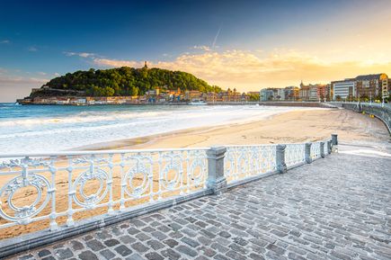 La Concha in San Sebastián, Spanien, mit goldfarbenem Sand, sanften Wellen, einer geschwungenen Promenade und der Stadtkulisse mit bewaldeten Hügeln im Hintergrund bei Sonnenuntergang.