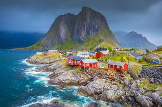 Felsige Küste am Meer in Norwegen mit roten Häusern auf den Felsen. Im Hintergrund ist ein großer steiniger Berg.