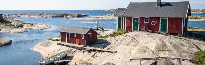 Ein kleines, rotes Holzhaus am Wasser, umgeben von Felsen und Bäumen. Boote liegen im ruhigen Wasser, und der Himmel ist blau.