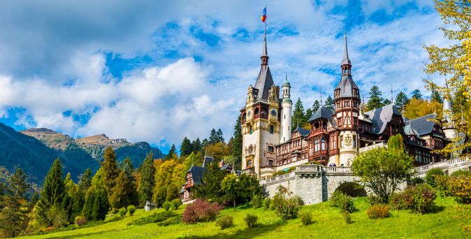 Blick auf das große Schloss Peles in Sinaia. Im Vordergrund viel Wiese und Bäume, etwas höher versetzt steht das Schloss und dahinter folgt wieder Wald, eine Bergkette und blauer Himmel.