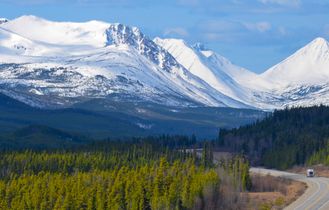 Ein Campervan fährt auf dem Alaska Highway im Yukon, Kanada, durch eine weite, bewaldete Landschaft, umgeben von Bergen unter einem bewölkten Himmel.
