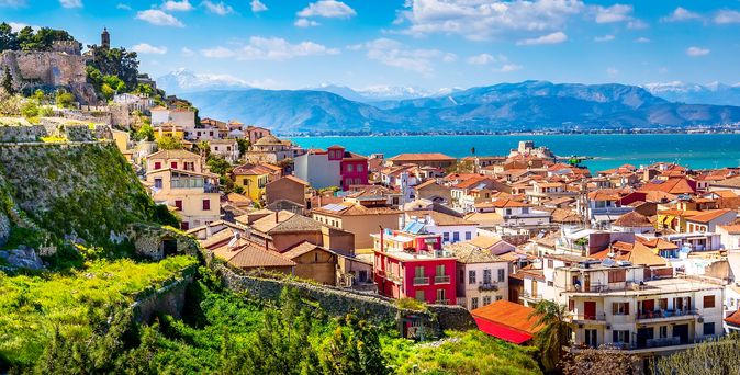 Blick über Nafplio mit seinen bunten Häusern, einem kleinem Strand und Hafen. In der Ferne ist eine Bergkette zu sehen. Aufgenommen bei strahlend blauem Himmel.