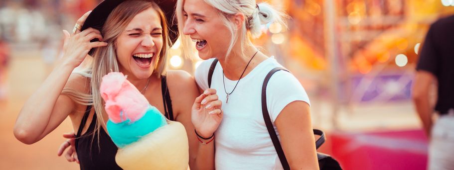 Zwei junge Frauen lachen und umarmen sich auf einem Jahrmarkt, während eine von ihnen bunte Zuckerwatte hält und die Lichter des Jahrmarkts im Hintergrund verschwimmen.