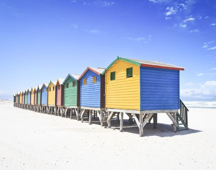 Eine Reihe von bunten Strandhütten in leuchtenden Farben am Muizenberg Beach in Kapstadt, Südafrika, mit dem weiten Sandstrand und dem Meer im Hintergrund.