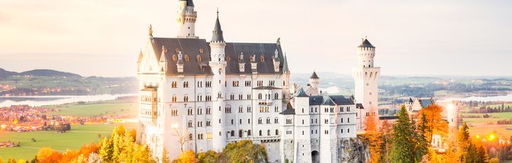 Ein malerisches Schloss oder eine Burg auf einem Hügel, umgeben von Bäumen in Herbstfarben und einem See, der die Landschaft widerspiegelt.