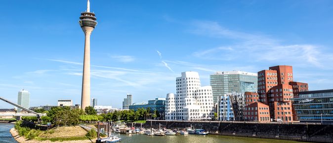 Blick auf den MedienHafen Düsseldorf mit dem Rheinturm und modernen Gebäuden am Flussufer unter blauem Himmel.