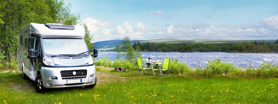 Ein silbernes Wohnmobil parkt auf einer blühenden Wiese an einem See, daneben ein Campingtisch mit Stühlen, bereit für eine Mahlzeit im Freien.