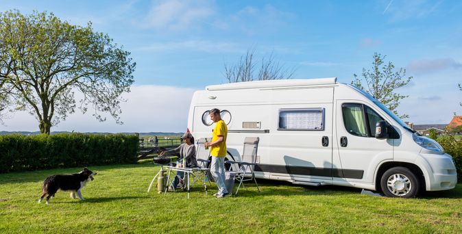 Camper steht auf die Wiese, davor sitzt eine Frau und steht ein Mann mit gelben Pulli. Vor ihnen steht der schwarze, kniehohe Hund. Eingezäunt, mit Bäumen umrandetes Grundstück. 