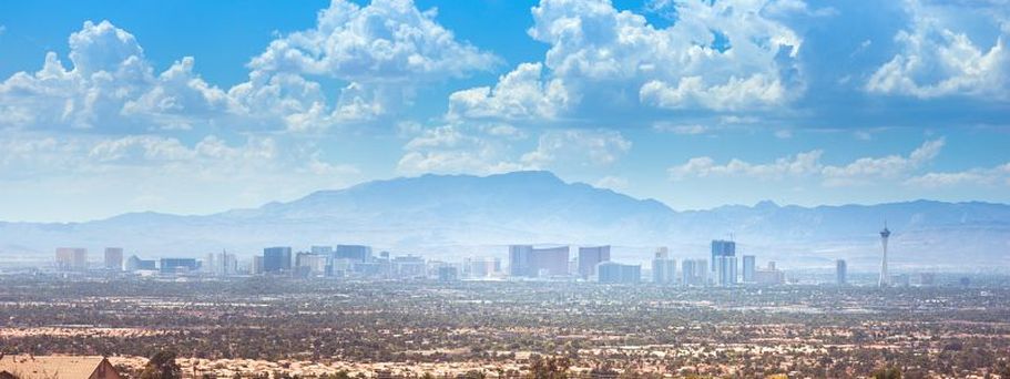 Panoramaaufnahme von Las Vegas mit Blick auf die Skyline des Strip und umliegende Vorstadtviertel vor einer Gebirgskulisse unter blauem Himmel.