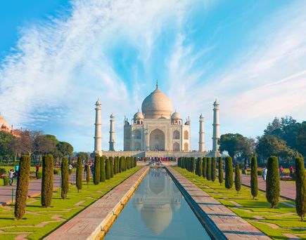 Taj Mahal in Indien. Blick von gegenüberliegender Stelle, davor der lange Pool, links und rechts Bäume, Wiese und Wege. Gegenüber liegt dann das imposante Gebäude und erstrahlt bei blauem Himmel.