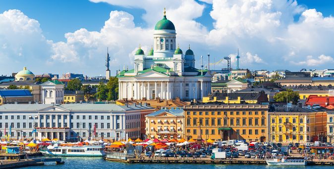 Blick auf den Hafen von Helsinki mit buntem Markttreiben am Ufer und dem weißen Dom von Helsinki im Hintergrund. Der Himmel ist blau mit großen, weißen Wolken.