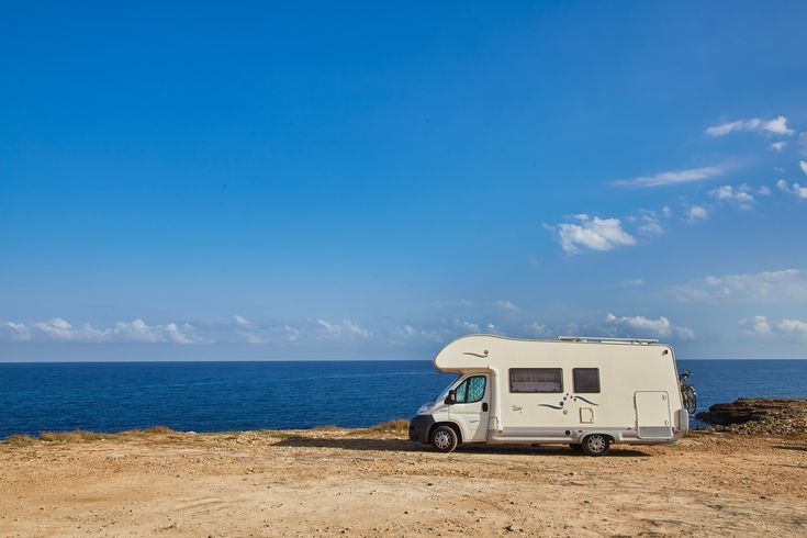 Ein Wohnmobil steht an einer trockenen Küste, das Meer und strahlend blauer Himmel im Hintergrund.
