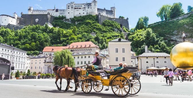 Eine Kutsche im Vordergrund, nähe Marktplatz. Daher ein Hügel mit der Festung Hohensalzburg.