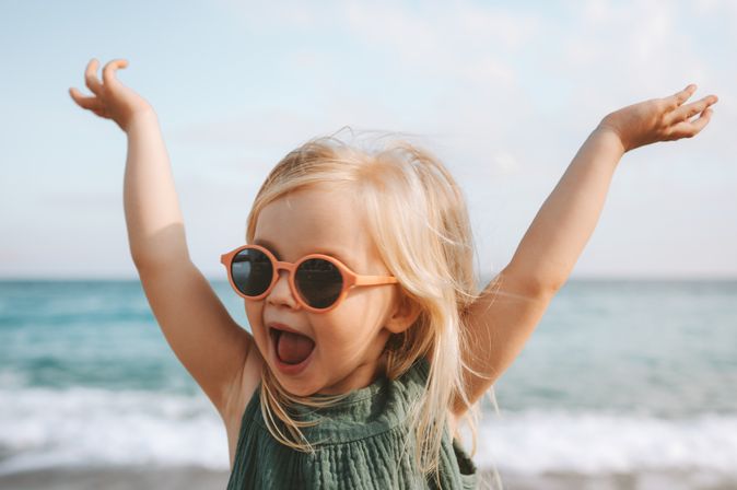 Kleines Kind mit Sonnenbrille steht am Meer und streckt die Arme nach oben