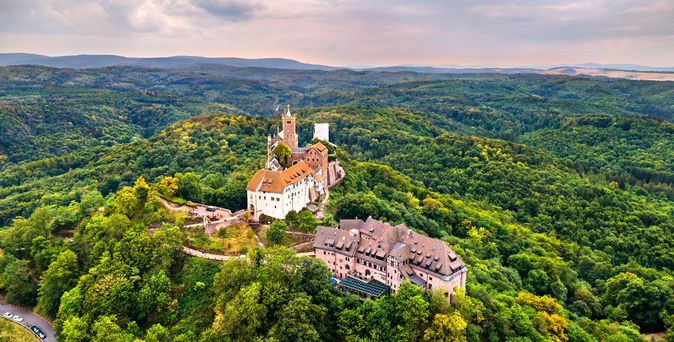 Ein majestätisches Luftbild der Wartburg, die auf einem bewaldeten Hügel thront und von einem weiten Waldgebiet umgeben ist, unter einem leicht bewölkten Himmel.