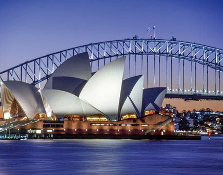 Sydney Opera House mit seinen beleuchteten weißen Segeldächern bei Sonnenuntergang, im Hintergrund die Sydney Harbour Bridge über dem Hafen.