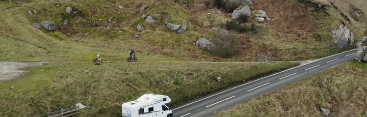 Ein weißes Wohnmobil fährt auf einer kurvenreichen Straße durch eine weite, grasbewachsene Landschaft mit Seen und Bergen im Hintergrund. Der Himmel ist bewölkt und spiegelt sich im Wasser.