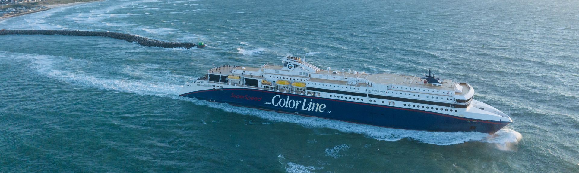 Ein blau-weißes Fährschiff mit der Aufschrift "ANEK LINES" auf der Seite bewegt sich auf dem Wasser, mit einer großen Welle hinter sich, unter einem hellen Himmel.