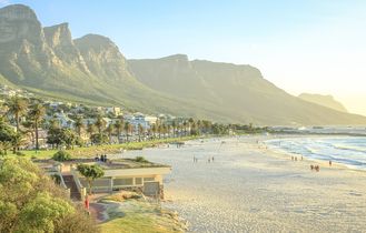 Eine atemberaubende Panoramaansicht der Camps Bay Beach in Kapstadt, Südafrika, mit dem weißen Sandstrand, dem türkisfarbenen Wasser und den majestätischen Zwölf Apostel Bergen im Hintergrund.