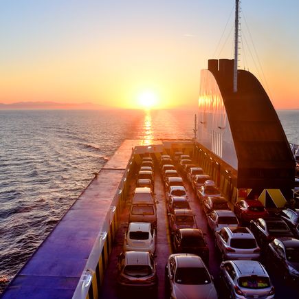 Eine Person steht am Heck eines Schiffes und blickt auf den Sonnenuntergang über dem Meer. Die orangefarbenen und rosa Farbtöne spiegeln sich auf dem Wasser.